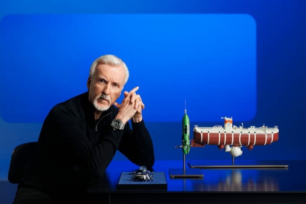 James Cameron, une Oyster Perpetual Deepsea Challenge au poignet, posant avec une maquette du bathyscape Trieste, son submersible DEEPSEA CHALLENGER (au centre) et les deux montres expérimentales fixées aux appareils pendant ses plongées dans la fosse des Mariannes (à gauche) - Respectivement la Deep Sea Special, (en arrière plan) et la Rolex Deepsea Challenge (devant).