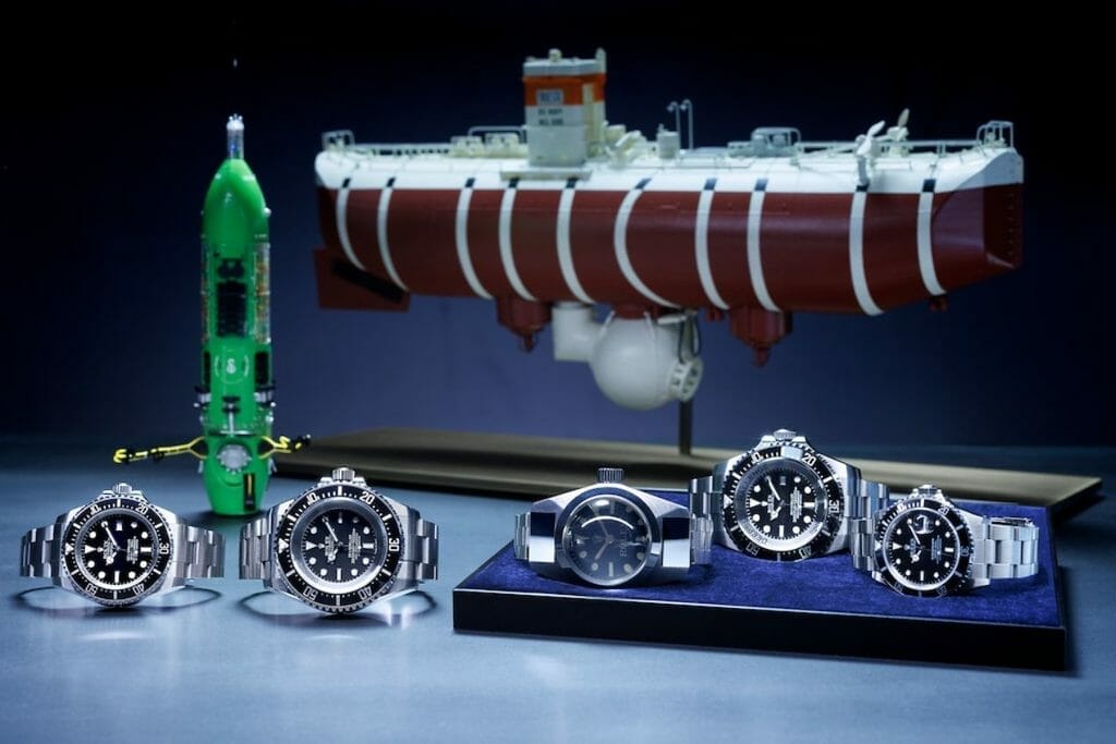 Premier plan, de gauche à droite : Oyster Perpetual Rolex Deepsea (2008), Oyster Perpetual Deepsea Challenge (2022), Deep Sea Special (1960), Rolex Deepsea Challenge (2012) et Oyster Perpetual Submariner (1986). Arrière-plan de gauche à droite : maquettes du submersible DEEPSEA CHALLENGER et du bathyscaphe Trieste. 
